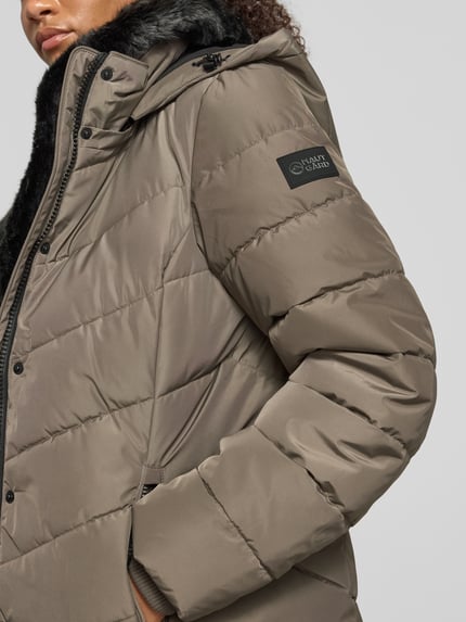 Frauenmodel trägt Regular Fit Jacke in Stepp-Optik von NAUTGÅRD in Taupe mit Kunstfellbesatz und Eingrifftaschen, Nahaufnahme Oberkörper. Rückenlänge 68 cm, Schulterbreite 43 cm, Brustweite 47 cm, Ärmellänge 66 cm.