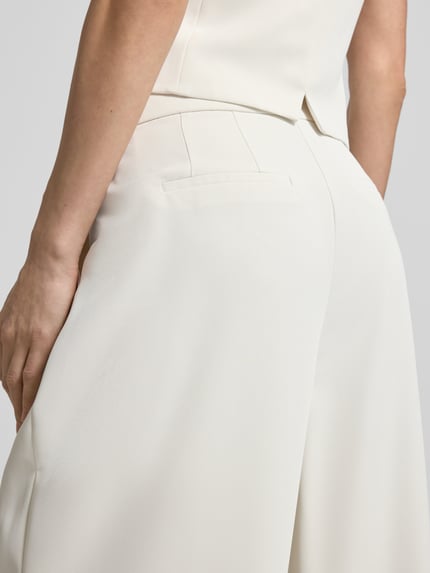 Kobiece modelka nosi Wide Leg Stoffhose mit Raffungen od Christian Berg Cocktail, elastyczna mieszanka materiałów, kremowy kolor, długość wewnętrzna nogawki 82 cm, szerokość w rozmiarze 36: 38 cm, zapięcie na haftkę i zamek błyskawiczny, marszczenia, widok z tyłu, zbliżenie na biodra.