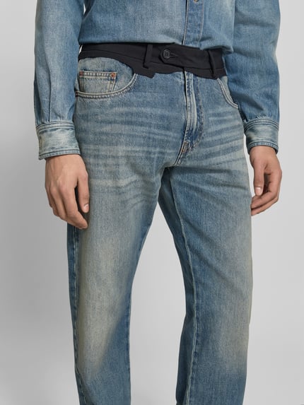 Męski model nosi Straight Fit Jeans mit kontrastivem Bund od MM6 Maison Margiela, jasnoniebieski denim, 5 kieszeni, zapięcie na guzik i zamek, bawełna, szerokość w rozmiarze 32: 42 cm, długość nogawki: 87 cm, zdjęcie z przodu, zbliżenie na górną część spodni.