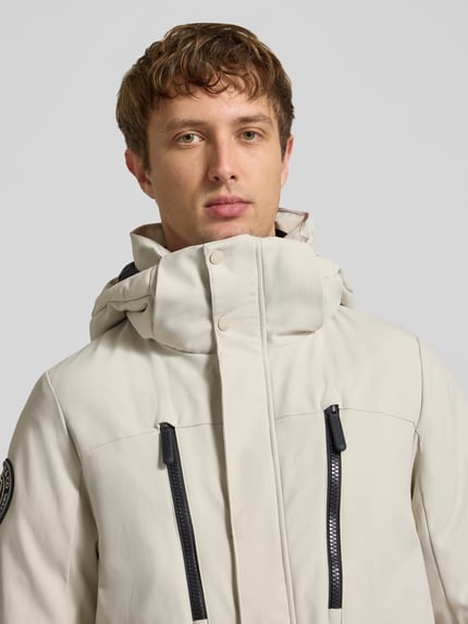 Männermodel trägt Parka mit Kapuze und kieszenie z patką von Superdry, krój regular fit, jasny beż, zamek błyskawiczny, Nahaufnahme Oberkörper. Długość tyłu w rozmiarze M: 80 cm, szerokość ramion w rozmiarze M: 48 cm, szerokość klatki piersiowej w rozmiarze M: 61 cm, długość rękawów w rozmiarze M: 73 cm.