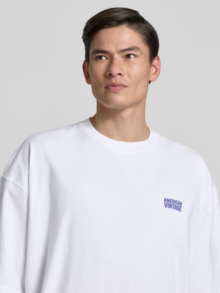 Männermodel trägt weißes Herren T-Shirt mit Logo-Print von American Vintage, Reine Baumwolle, Loose-Fit, Nahaufnahme Oberkörper. Modell 'GIXY', Rundhalsausschnitt, Rückenlänge 72 cm bei Größe M/L.
