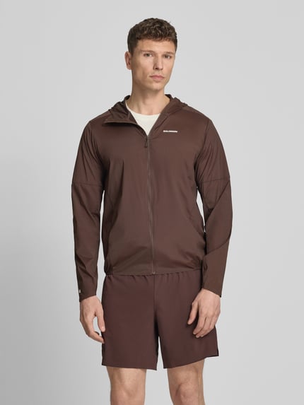 Männermodel trägt Windbreaker mit Kapuze und Logo Modell 'SENSE AERO' von Salomon aus Material-Mix mit Stretch-Anteil, Regular-Fit, Reißverschluss, Rückenlänge 71 cm, Schulterbreite 44 cm, Brustweite 41 cm, Ärmellänge 67 cm in Braun, Ganzkörperansicht von vorne.