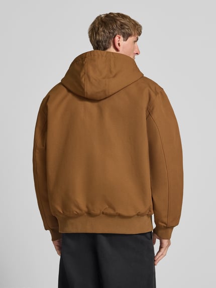 Männermodel trägt Jacke mit Label-Patch und Känguru-Tasche von Carhartt Work In Progress in Braun mit Kapuze, Rückansicht, Ganzkörperansicht Rückansicht. Reine Baumwolle, Regular Fit, gerippte Abschlüsse, sichtbare Textur.
