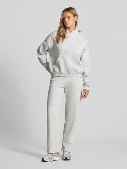 Frauenmodel trägt Relaxed Fit Hoodie aus Baumwoll-Mix Modell 'Iconic' von Gina Tricot in Hellgrau mit Känguru-Tasche, elastische Abschlüsse, Ganzkörperansicht von vorne.