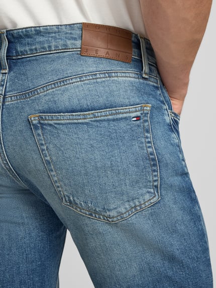 Männermodel trägt Slim Fit Jeans aus Baumwoll-Mix Modell 'SCANTON SLIM' von Tommy Jeans in Denim-Optik mit Logo-Stitching, Detailaufnahme Rückansicht. 5-Pocket-Design, sichtbare strukturierte Textur und Kontrastnähte, Kontext: Herren Jeans von Tommy Jeans, Passform: Slim Fit, Applikation: Logo-Stitching, Verschluss: Knopf und verdeckter Reißverschluss, Material: Baumwoll-Mix.
