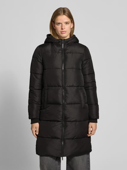 Frauenmodel trägt Puffermantel mit Kapuze von PIECES in Schwarz, Longline-Form, Reißverschluss, Kapuze, seitliche Taschen, Ganzkörperansicht von vorne.