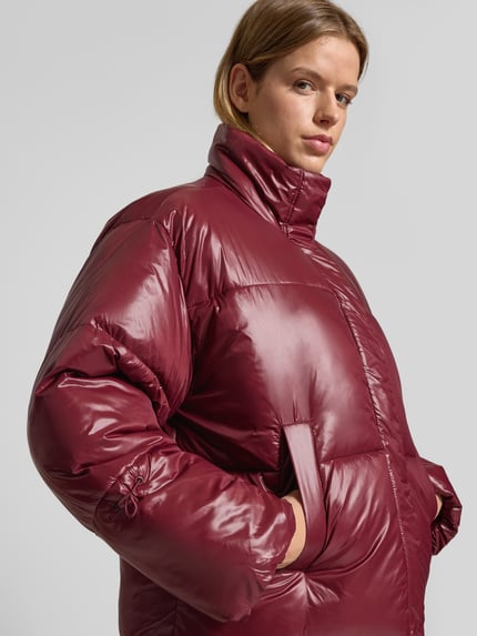 Kobiece modelka nosi kurtkę pikowaną z stójką Noisy May, model 'ALLIE', kolor bordowy, krój relaxed fit, wpuszczane kieszenie, zamek błyskawiczny zakryty, gładka faktura, długość tyłu 66 cm w rozmiarze S, szerokość ramion 56 cm, szerokość klatki piersiowej 66 cm, długość rękawów 56 cm, całkowity widok z przodu.