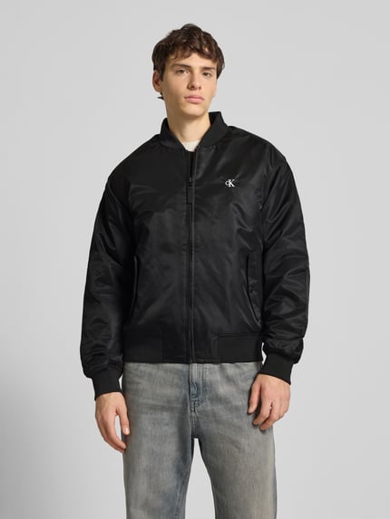Männermodel trägt Bomberjacke mit Label-Stitching von Calvin Klein Jeans in Schwarz, Regular Fit, mit Stehkragen und Taschen, Ganzkörperansicht von vorne.