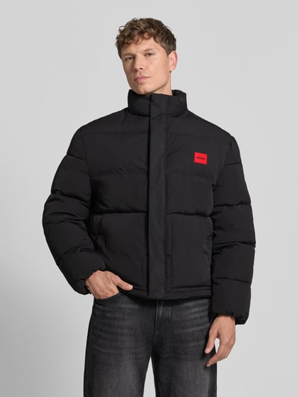 Männermodel trägt Daunenjacke mit Logo-Patch von HUGO in Unifarben mit Stehkragen, Reißverschluss und Taschen, Ganzkörperansicht von vorne