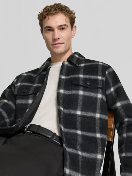 Männermodel trägt Hemdjacke mit Druckknopfleiste von BOSS Orange im schwarzen Karo-Allover-Muster, Nahaufnahme Oberkörper.