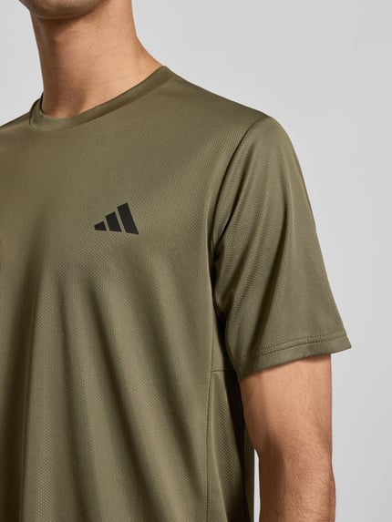 Männermodel trägt T-Shirt mit Label-Print von adidas Training in Oliv, Lockerer-Schnitt, gerippter Rundhalsausschnitt, Nahaufnahme Oberkörper. Rückenlänge bei Größe M: 70 cm.