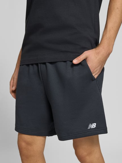 Männermodel trägt schwarze Sweatshorts aus Baumwoll-Mix Modell 'FRENCH' von New Balance, elastischer Bund, Logo-Prägung, Nahaufnahme Oberkörper, Kontext: Herren Sweatshorts, gerade Passform, Gesäßtasche, Bundweite 40 cm, Innenbeinlänge 17 cm.
