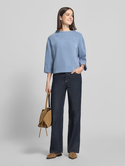 Frauenmodel trägt Relaxed Fit Strickpullover mit Stehkragen von ZABAIONE in Strick-Optik, Logo-Emblem, 3/4-Arm, Ärmelschlitze, überschnittene Schultern, hellblau, kombiniert mit dunkelblauer Hose und beigefarbener Tasche, Ganzkörperansicht von vorne.