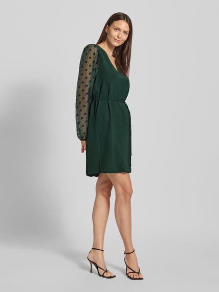Frauenmodel trägt fließendes Cocktailkleid mit semitransparenten Ballonärmeln Modell 'ALVA' von Vero Moda in dunklem Unifarben, Mini-Länge, V-Ausschnitt, Crinkle Materialverarbeitung, Optik: Unifarben/semitransparent, Passform: fließend, Ganzkörperansicht von vorne.