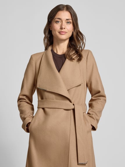 Vrouwenmodel draagt lange jas Model 'ROSE' van Ted Baker in camel, met opstaande kraag, knoopsluiting, labeldetail, strikceintuur, Nahaufnahme Oberkörper. Ruglengte maat 36: 105 cm, schouderbreedte: 40 cm, borstwijdte: 45 cm, mouwlengte: 63 cm.