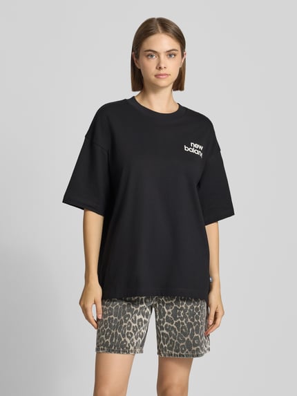 Frauenmodel trägt schwarzes Relaxed Fit T-Shirt mit Label-Print Modell 'INTELLIGENT' von New Balance, reine Baumwolle, Rundhalsausschnitt, Ganzkörperansicht von vorne.