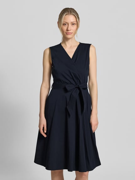 Kobiece model nosi sukienkę 'Kleid mit V-Ausschnitt in Wickel-Optik' od More & More, mieszanka bawełny, taliowany krój, dekolt w serek, kopertowy styl, bez rękawów, odpinany pasek w talii, długość tyłu w rozmiarze 36: 100 cm, szerokość w rozmiarze 36: 35 cm, kolor granatowy, jednolita faktura, widok całej sylwetki z przodu.