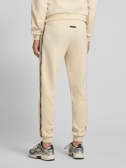Męski model nosi spodnie dresowe Sweatpants aus Baumwoll-Mix od EA7 Emporio Armani, mieszanka bawełny i elastanu, luźny krój, elastyczny pas z tunelem, zakończenia nogawek elastyczne, wpuszczane kieszenie, długość wewnętrzna nogawki w rozmiarze m: 78 cm, całkowity widok z tyłu.
