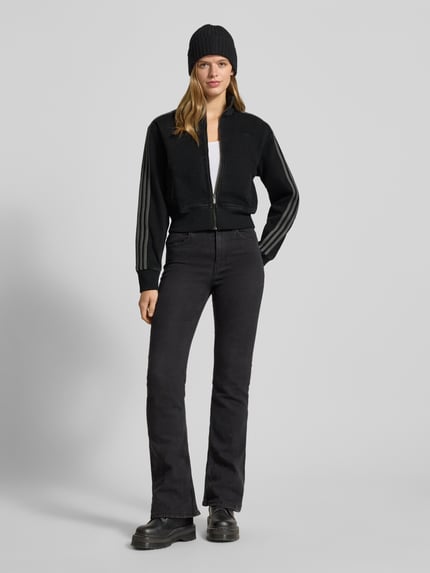 Kobiece model nosi Kurtka z zamkiem błyskawicznym od adidas Originals o kroju regular fit i skróconym kroju, czarny kolor, prążkowane wykończenia, stójka, wyhaftowane logo, wpuszczane kieszenie, długość tyłu 51 cm, szerokość ramion 45 cm, szerokość klatki piersiowej 41 cm, długość rękawów 62 cm; pełen widok z przodu.