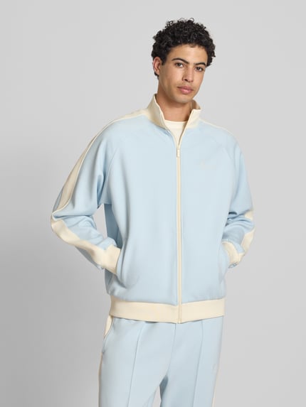 Männermodel trägt Regular Fit Sweatjacke mit Logo-Stitching von KARL KANI in Hellblau-Creme, Elastischer-Material-Mix, Stehkragen, Reißverschluss, Eingrifftaschen, elastische Abschlüsse, Rückenlänge 65 cm, Ärmellänge 74 cm, Ganzkörperansicht von vorne.