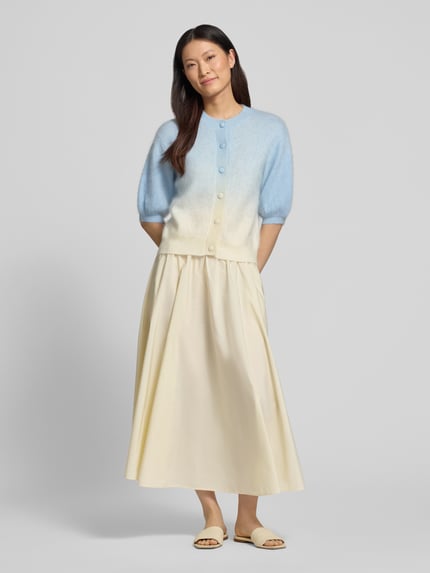 Frauenmodel trägt Strickjacke aus Kaschmir-Baumwoll-Mix mit 1/2-Arm von HERZENS in Blau und Creme, Regular-Fit, Rundhalsausschnitt, Knopfleiste, Puffärmel, kombiniert mit einem hellen, weit fallenden Rock; man erkennt weiche, flauschige Textur und sanfte Farbverläufe, Ganzkörperansicht von vorne. Rückenlänge bei Größe 36: 56 cm, Ärmellänge 26 cm.