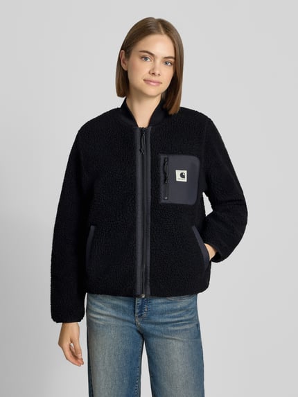 Kobiece modelka ma na sobie kurtkę z polaru z Futerko Carhartt Work In Progress, Model 'Janet', czarna, wpuszczane kieszenie, Brustasza, detal z logo, zamek błyskawiczny dwustronny, stójka, krój regular fit, widok całego ciała z przodu.