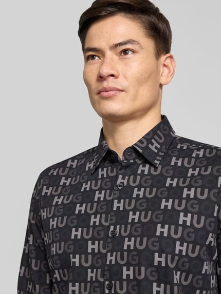 Męski model nosi Slim Fit Freizeithemd aus reiner Baumwolle Modell 'ERMO' von HUGO z czarnym wzorem logo, zbliżenie na górę ciała.