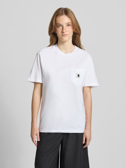 Kobiece modelka nosi damski t-shirt T-Shirt mit Brusttasche und Label-Detail von Carhartt Work In Progress, bawełna, krój regular fit, okrągły dekolt, brustasza, detal z logo, długość tyłu w rozmiarze S: 59 cm, biała gładka tkanina, przód na całej sylwetce, widok z przodu