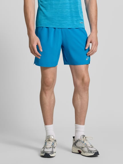 Männermodel trägt Regular Fit Shorts mit integriertem Slip Modell 'Core' von ASICS TIGER in Blau, Logo-Print, elastischer Bund, Ganzkörperansicht von vorne. Herren Shorts, Bundweite M: 38 cm, Innenbeinlänge M: 17 cm, glatte Textur.