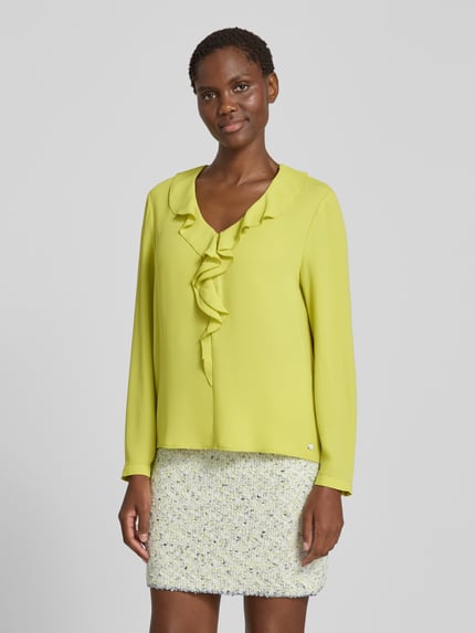 Kobiece model prezentuje Relaxed Fit Bluse mit Rüschenbesatz od Marc Cain, bluzka z obszyciem falbanami, dekoltem w serek, z długim rękawem, długość tyłu w rozmiarze 36: 58 cm, długość rękawów w rozmiarze 36: 61 cm, jasny żółty kolor, gładka tekstura, widok całej sylwetki z przodu.