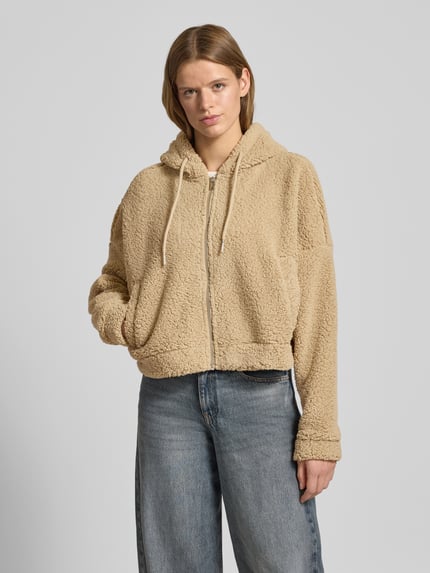 Kobiece modelka nosi Kurtka Cropped Jacke aus Teddyfell Model 'CUDDLE' od Noisy May w kolorze beżowym, luźny krój, futerko, kaptur, tunel, zamek błyskawiczny, wpuszczane kieszenie, długość tyłu 48 cm, szerokość ramion 79 cm, szerokość klatki piersiowej 77 cm, długość rękawów 48 cm, widok całej sylwetki z przodu.
