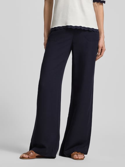 Kobieta nosi Wide Fit Stoffhose aus Viskose-mix mit Leinen-Anteil od Marc Cain, mieszanka wiskozy i lnu, detal z logo, kieszenie z wypustkami, wpuszczane kieszenie, szerokość w rozmiarze 36: 37 cm, długość wewnętrzna nogawki w rozmiarze 36: 82 cm, ciemnogranatowy kolor, gładka tekstura, spodnie materiałowe, Ganzkörperansicht z przodu.