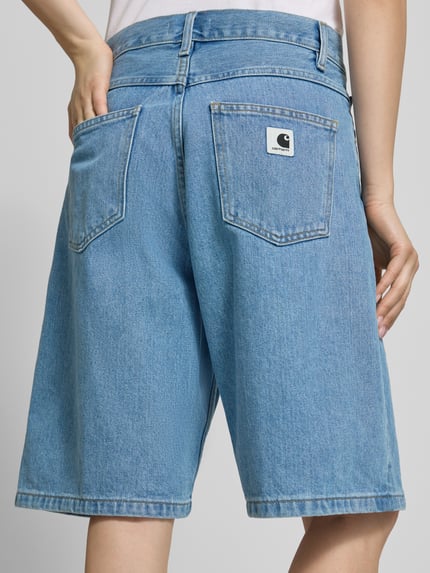 Frauenmodel trägt Relaxed Fit Jeans Shorts aus reiner Baumwolle Modell 'Brandon' von Carhartt Work In Progress, hellblauer Denim, 5-Pocket-Design, Label-Patch, Knopf-Reißverschluss, Ganzkörperansicht Rückansicht.