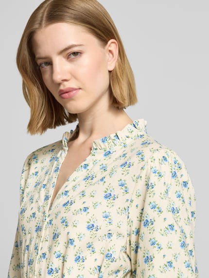Vrouwenmodel draagt knielang jurk uit linnenmix model 'Solfried' van MSCH Copenhagen, getailleerd, met viscose, halve mouw, opstaande kraag met ruches, ruglengte maat S 96 cm, borstwijdte maat S 84 cm, mouwlengte maat S 37 cm, crèmekleurig met blauwe bloemenprint, textuur glad. Nahaufnahme bovenlichaam