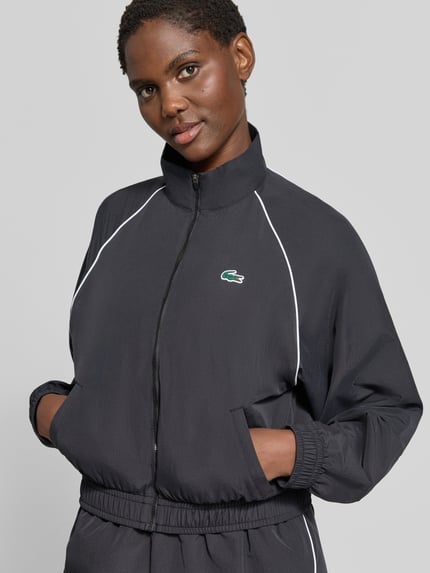 Frauenmodel trägt Relaxed Fit Trainingsjacke mit Logo-Patch von Lacoste in Schwarz mit weißem Piping, glatte Struktur, Nahaufnahme Oberkörper, Damen Trainingsjacke von Lacoste Sport.