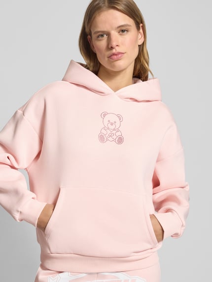 Frauenmodel trägt Hoodie mit Kapuze von Review in zartem Rosa, Ziersteinbesatz, Känguru-Tasche, lockerer Schnitt, Nahaufnahme Oberkörper.