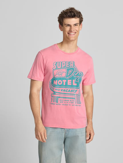 Männermodel trägt Loose Fit T-Shirt mit Label-Print Modell 'Roadside Trade' von Superdry in Rosa und Blau, reine Baumwolle, Ganzkörperansicht von vorne.