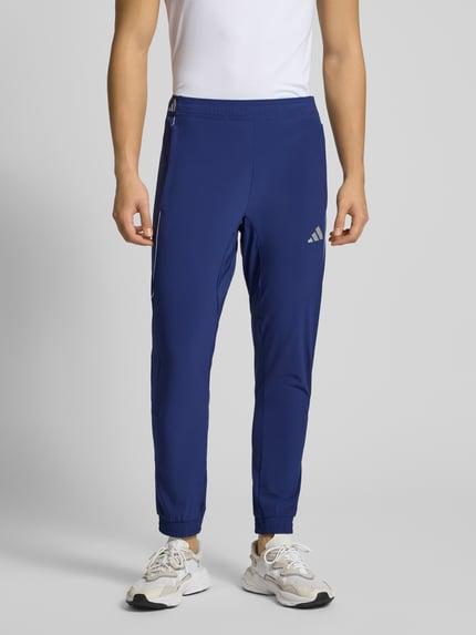 Männermodel draagt Slim Fit Sweatpants met Logoprint model 'Tech' van adidas Training in blauwe elastische materiaalmix, elastische band en boordjes, binnenbeenlengte maat M 75 cm, Ganzkörperansicht van voren.