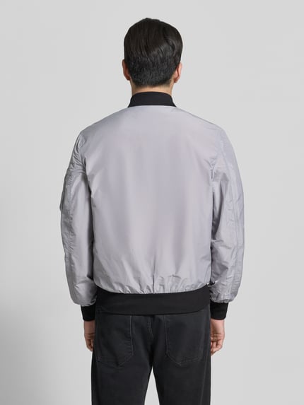 Männermodel trägt Bomberjacke mit Label-Patch und Eingrifftaschen von Original Bombers, grauer Farbton, Gerade geschnitten, Rückenansicht, Kontext: Herren Bomberjacke, Reißverschluss, Reißverschlusstasche am Ärmel, Druckknopfverschluss, Gerippte-Abschlüsse, Rückenlänge Größe M 67 cm, Schulterbreite 43 cm, Brustweite 53 cm, Ärmellänge 68 cm.