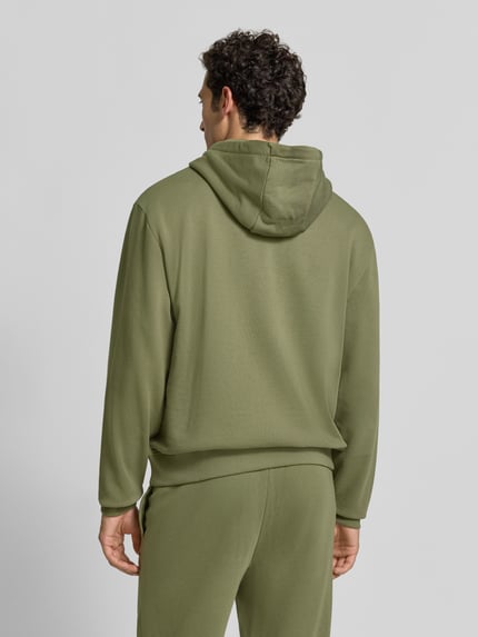 Männermodel trägt Relaxed Fit Hoodie aus reiner Baumwolle Modell 'Dapo' von HUGO in Olivgrün, glatte Textur, Ganzkörperansicht Rückansicht. Herren Hoodie von HUGO.