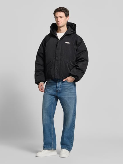 Männermodel trägt Relaxed Fit Bomberjacke mit Label-Patch Modell 'L’ATELIER' von RETERNITY in Schwarz, gerade Jeans, weiße Sneaker, Ganzkörperansicht von vorne. Rückenlänge, Schulterbreite, Brustweite und Ärmellänge gemäß Größe M.