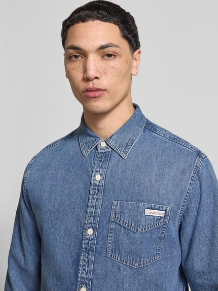 Mężczyznamodel nosi Regular Fit Jeanshemd mit Brusttasche und Label-Detail von Calvin Klein Jeans z bawełny, kołnierzyk typu kent, listwa guzikowa, detal z logo, kieszeń na piersi, długość tyłu w rozmiarze M 76 cm, szerokość ramion 47 cm, długość rękawów 63 cm, niebieski denim, prosty krój, tekstura jeansu. Zbliżenie na górną część ciała.