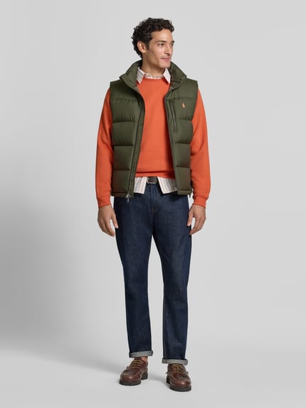 Männermodel trägt Regular Fit Weste mit Logo-Stitching von Polo Ralph Lauren in Oliv, kombiniert mit orangenem Pullover, Ganzkörperansicht von vorne.