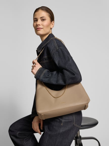 Frauenmodel trägt Handtasche mit Label-Detail von CK Calvin Klein in Beige, strukturierte Oberfläche, goldfarbener Kettenriemen, Nahaufnahme Oberkörper. Hauptfach mit Reißverschluss, Innenfach, Maße: 24x27x11 cm, Schulterriemen 68 cm.