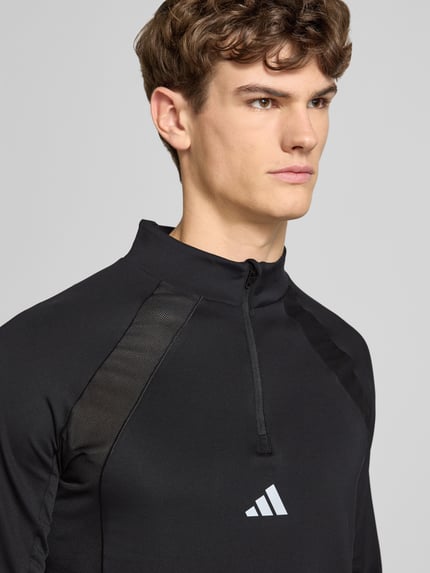 Männermodel trägt Sweatshirt mit Stehkragen von adidas Training, czarny materiał, krój slim fit, krótki zamek, nadruk z logo, Nahaufnahme Oberkörper.