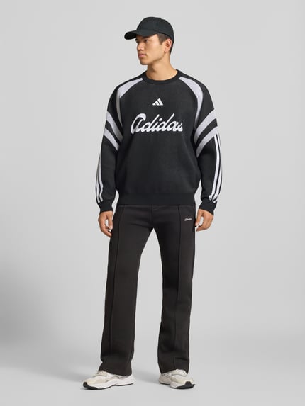 Männermodel trägt pullover met grote logo-opschrift van adidas Sportswear, meerkleurig, gebreide look, motiefprint, gestreept, logoprint, contraststrepen, ronde hals, lange mouw, mouw- en rompzoom geribd, vooraanzicht van volledig lichaam
