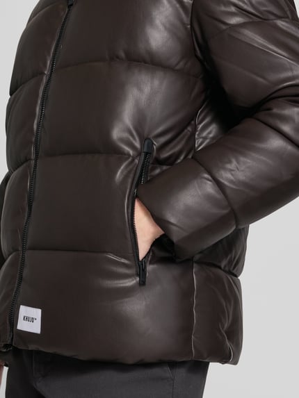 Männermodel trägt kurtka pikowana z kapturem Modell 'COEN 2' von khujo, krój regular fit, zamek błyskawiczny, detal z logo, kolor ciemny brąz, Długość tyłu w rozmiarze M: 72 cm, szerokość ramion w rozmiarze M: 55 cm, szerokość klatki piersiowej w rozmiarze M: 61 cm, Długość rękawów w rozmiarze M: 62 cm, detailaufnahme.