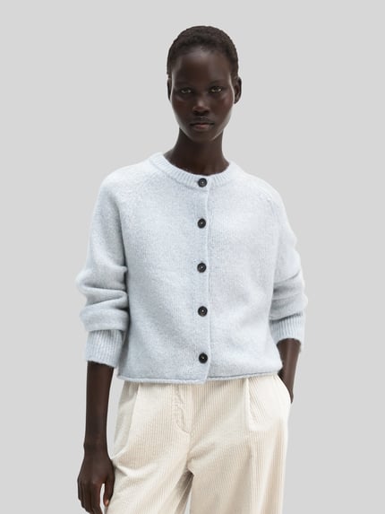 Frauenmodel trägt Strickjacke aus Alpaka Wolle mit Rollsaum von Marc O'Polo, hellblau-meliert, strukturierte Strickoptik, Baumwollmix, Raglanärmel, Rundhals, durchgehende Knopfleiste, Cropped-Länge, Ganzkörperansicht von vorne.