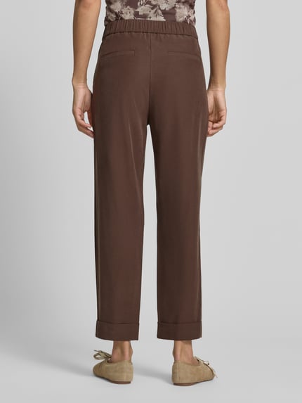 Frauenmodel trägt Stoffhose mit francuskimi wpuszczanymi kieszeniami od Jake*s Collection, krój straight fit, elastyczny pas, szerokość w rozmiarze 38: 36 cm, długość wewnętrzna nogawki 68 cm, kolor brązowy, gładka tekstura, kieszenie z wypustkami z tyłu, nogawki z zakładką, widoczne przeszycia, całkowity widok z tyłu.