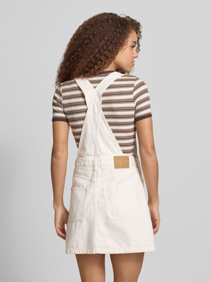 Frauenmodel trägt Leicht ausgestelltes Jeanskleid aus reiner Baumwolle von Tommy Jeans in heller Denim-Optik mit Brusttasche, Mini-Länge, Label-Stitching-Applikation und Latzträgern, Ganzkörperansicht Rückansicht.